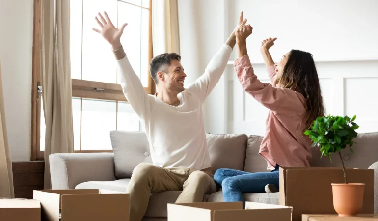 a man and woman happily raising their hands while sitting on a couch with cardboard boxes in front of them